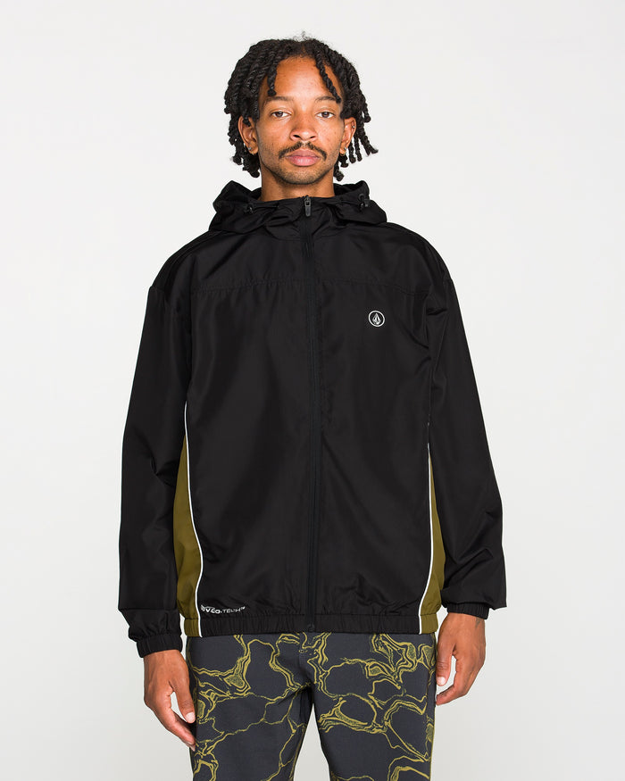 A person stands facing forward against a plain white background, wearing the Volcom Men's Wavestone Jacket in black, featuring a mesh-lined hood and white piping, paired with patterned black pants.