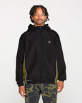 A person stands facing forward against a plain white background, wearing the Volcom Men's Wavestone Jacket in black, featuring a mesh-lined hood and white piping, paired with patterned black pants.