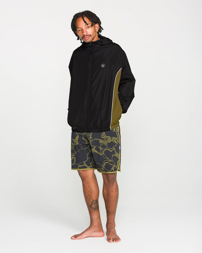 A man stands barefoot in front of a plain white background, wearing the Volcom Men's Wavestone Jacket—featuring a mesh-lined hood and olive green sides—paired with patterned black and yellow shorts.