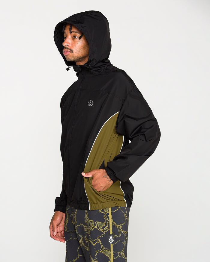 A man wearing the Volcom Men's Wavestone Jacket in black with olive green accents and a mesh-lined hood is paired with matching pants featuring a green lightning pattern, standing facing left against a plain white background.
