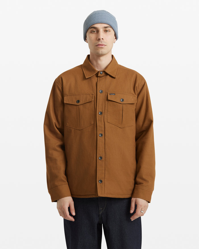 A person wearing a light blue knit beanie, dark jeans, and the Volcom Men's Westmead Jacket—a brown button-up with chest pockets—stands facing forward against a plain white background.