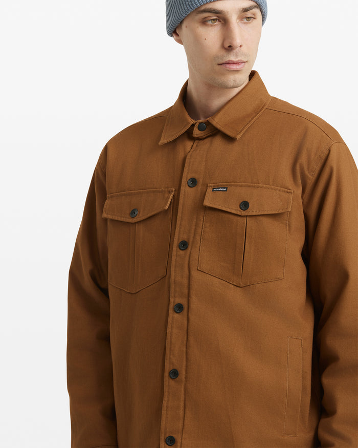 A man wearing a light blue knit beanie and the Volcom Men's Westmead Jacket, a brown button-up with two chest pockets, looks off to the side against a plain white background.