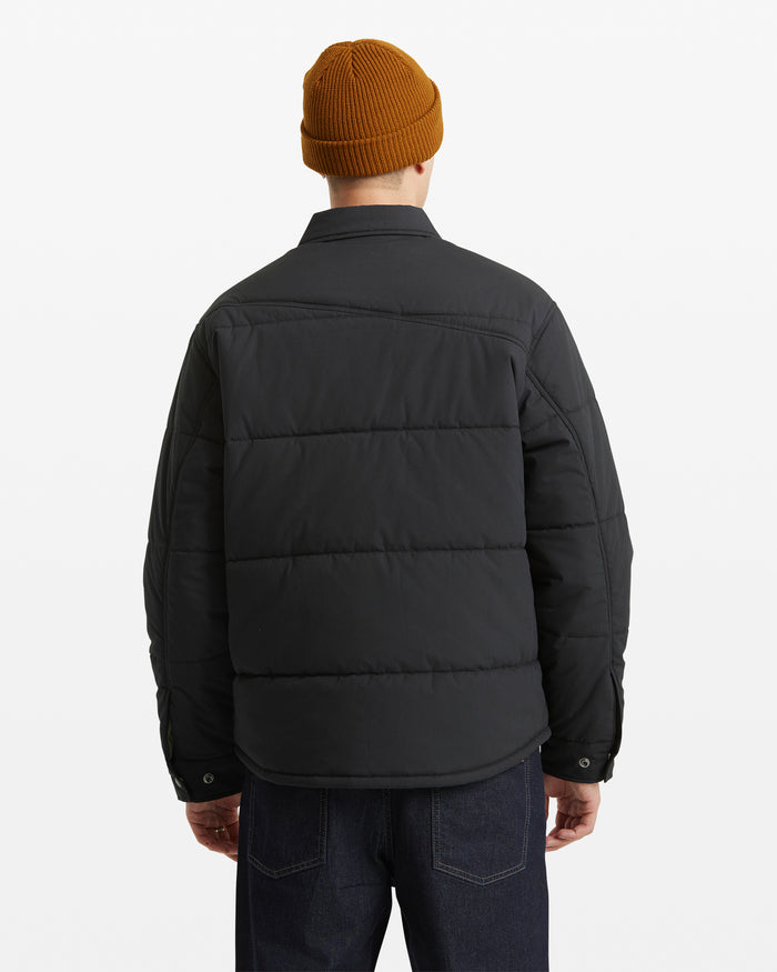 A person stands facing away against a plain white background, wearing the Volcom Men's Westmead Quilted Jacket in black, paired with a mustard knit beanie and dark jeans.