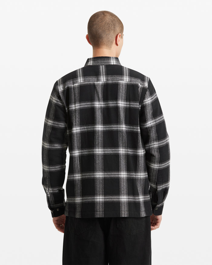 A person with short hair is shown from the back against a plain white background, wearing the Volcom Men's Wilderstone Long Sleeve in a black and white plaid pattern with black pants.