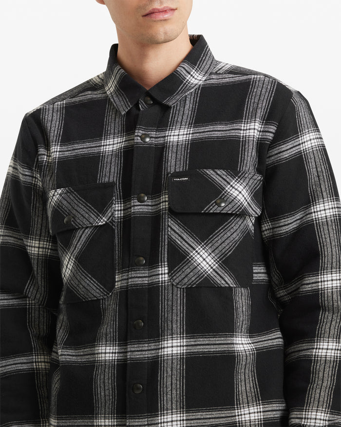 A man is shown from shoulders to waist against a plain white background, wearing the Volcom Men's Wilderstone Long Sleeve shirt—a black and white plaid cotton woven top with two chest pockets.