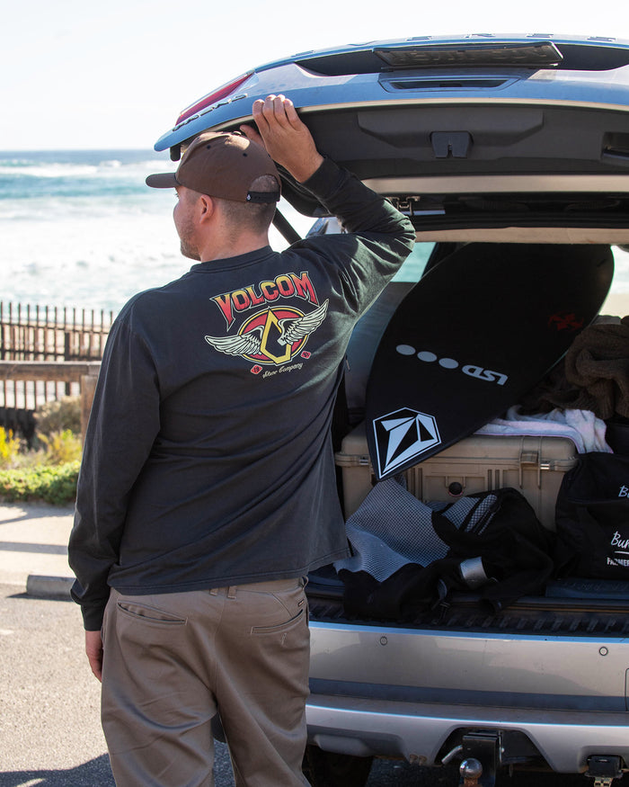 A person in a black, 100% cotton Volcom Men's Wing It Long Sleeve Tee and cap stands by an open car trunk of surf gear, holding the door. The ocean and a wooden fence are visible in the background.