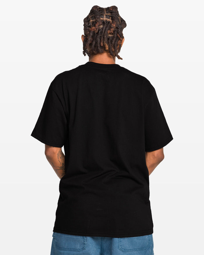 A person with short dreadlocks is seen from behind, wearing the Volcom Men’s Workwear Certifico Short Sleeve Tee in black—crafted from premium cotton for a classic fit—and blue denim shorts, standing against a plain white background. A woven label patch adds a subtle detail to the T-shirt’s design.