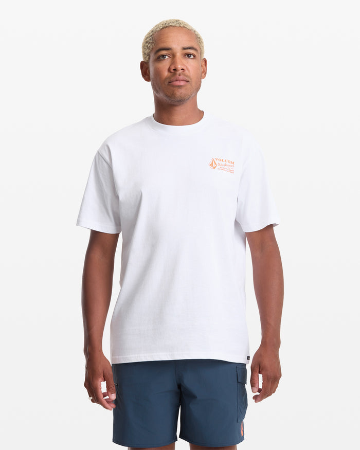 A person with short, light blonde hair wears the Volcom Men's Workwear Short Sleeve Tee in white—featuring a small orange logo and text on the upper left chest—and blue shorts, standing against a white background.