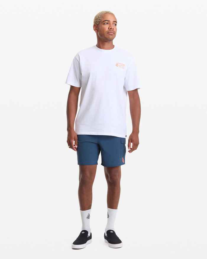 A person stands facing forward against a plain white background wearing a Volcom Men's Workwear Short Sleeve Tee in white, paired with blue shorts, white socks, and black slip-on shoes.