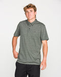 A young man with light brown hair wearing the Volcom Men's Wowzer Polo Short Sleeve Shirt in dark grey and black pants stands against a plain white background.