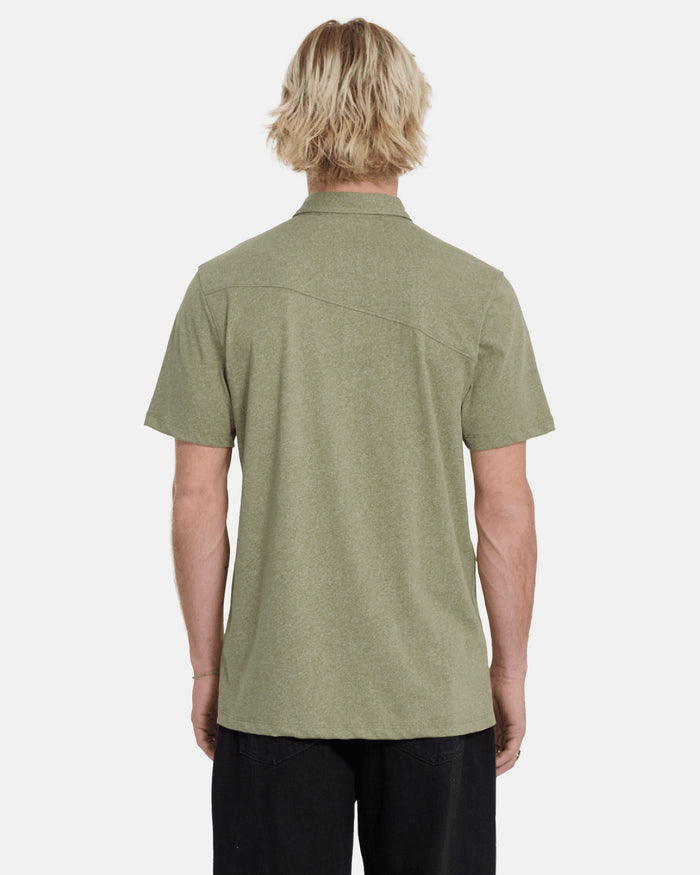 A person with light blonde, wavy hair is seen from behind wearing the Volcom Men's Wowzer Polo Short Sleeve Shirt in olive green and black pants against a plain, light background.