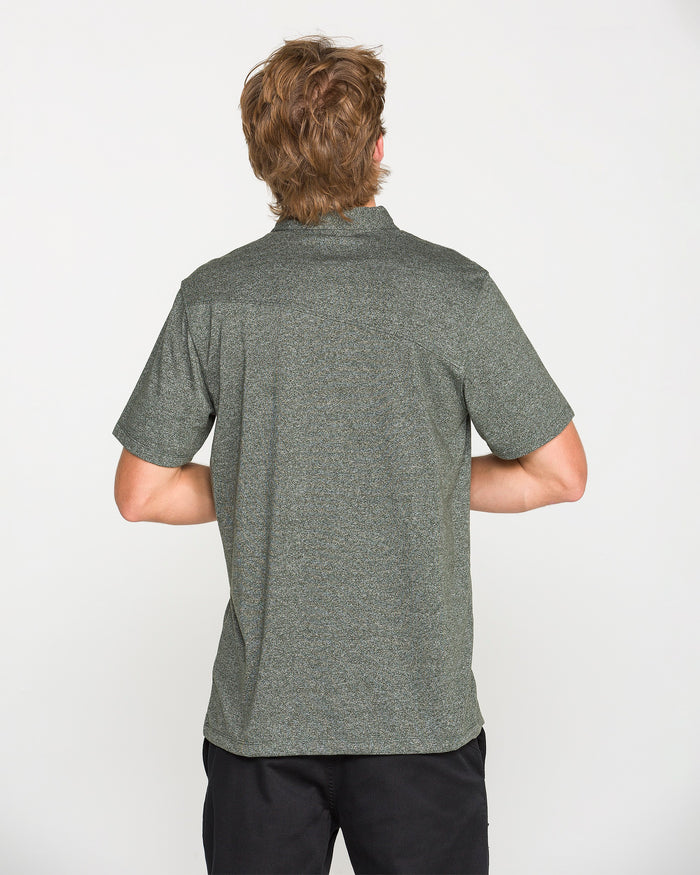 A person with short, light brown hair is standing facing away, wearing a Volcom Men's Wowzer Polo Short Sleeve Shirt in rosin green and black pants against a plain white background.