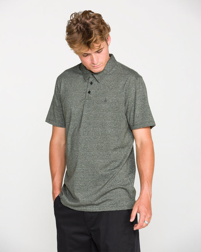 A man with light brown hair stands against a plain white background, looking down with his left hand in his pocket, wearing the Volcom Men's Wowzer Polo Short Sleeve Shirt in dark gray and black pants.