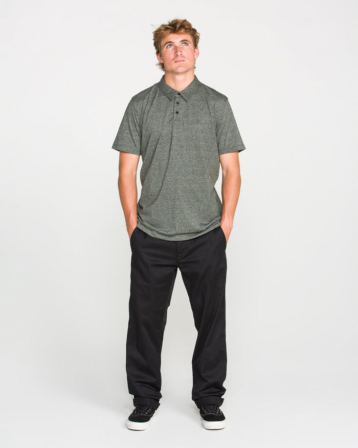 A man wearing the Volcom Men's Wowzer Polo Short Sleeve Shirt in gray, paired with black pants and black sneakers, stands with his hands in his pockets, looking upward against a plain white background.