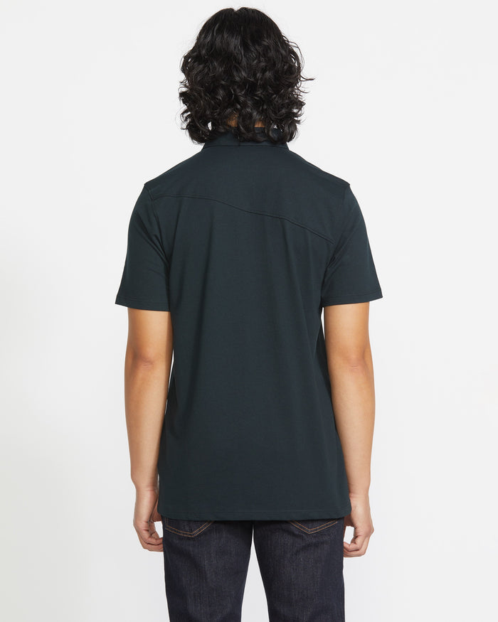 A person with shoulder-length wavy dark hair, wearing a Volcom Men's Wowzer Polo Short Sleeve Shirt in a dark cotton-poly blend and dark jeans, stands with their back to the camera against a plain white background.
