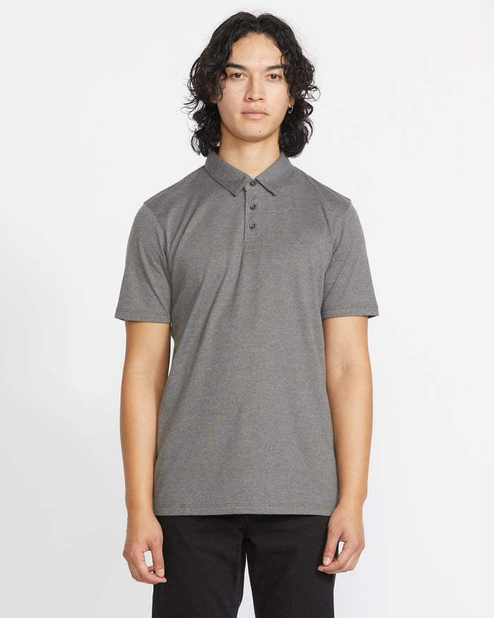 A person with medium-length curly hair wears the Volcom Men's Wowzer Polo Short Sleeve Shirt in gray, made from a cotton-poly blend with chest Stone embroidery, paired with black pants, standing against a white background facing forward.