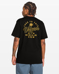 A person with braided hair faces away, wearing the Volcom Men's Wrecking Short Sleeve Tee—a classic-fit black shirt made from 100% cotton with yellow "True to This Volcom Stone Co Wrecking Crew" graphic text—paired with blue shorts on a white background.