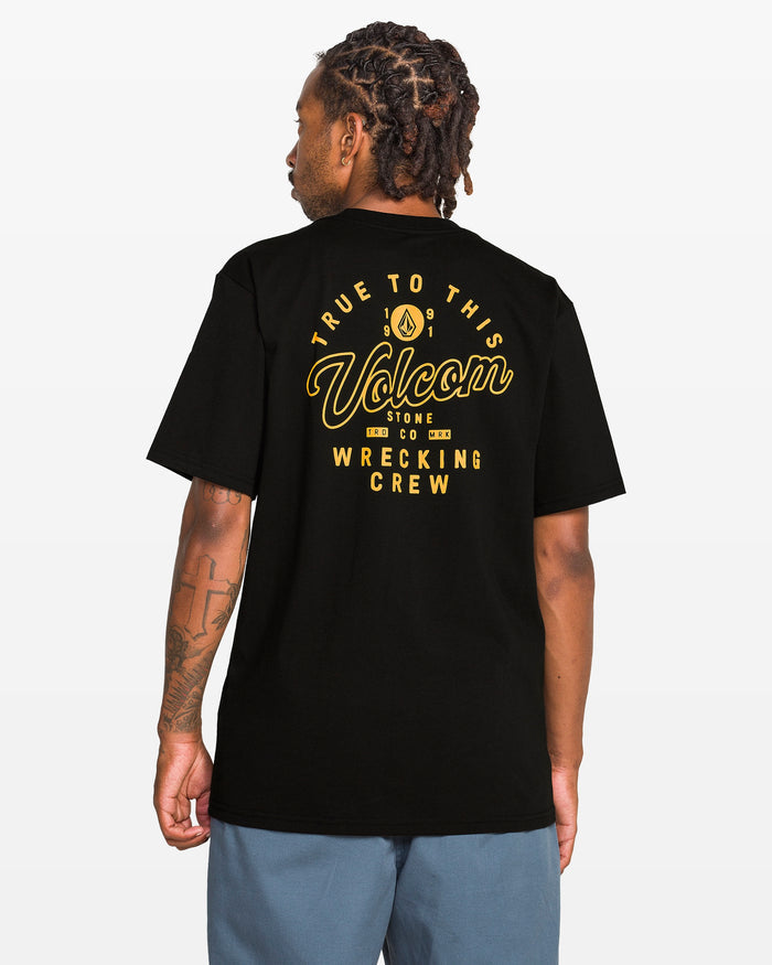 A person with braided hair faces away, wearing the Volcom Men's Wrecking Short Sleeve Tee—a classic-fit black shirt made from 100% cotton with yellow "True to This Volcom Stone Co Wrecking Crew" graphic text—paired with blue shorts on a white background.