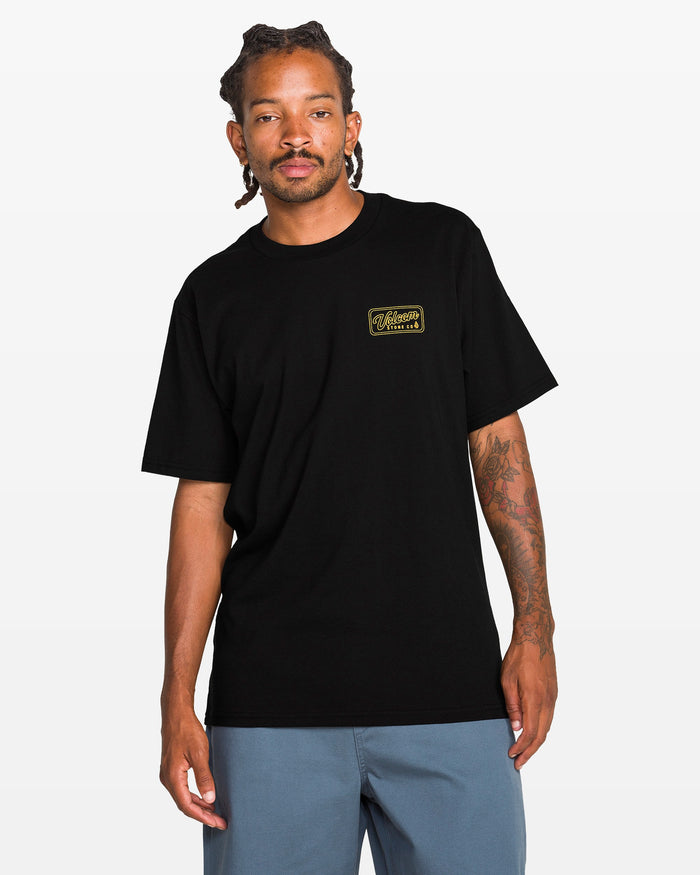 A man with braided hair and tattoos on his right arm wears the Volcom Men's Wrecking Short Sleeve Tee, a classic fit black 100% cotton shirt with a small yellow logo on the left chest, and blue shorts, against a plain white background.