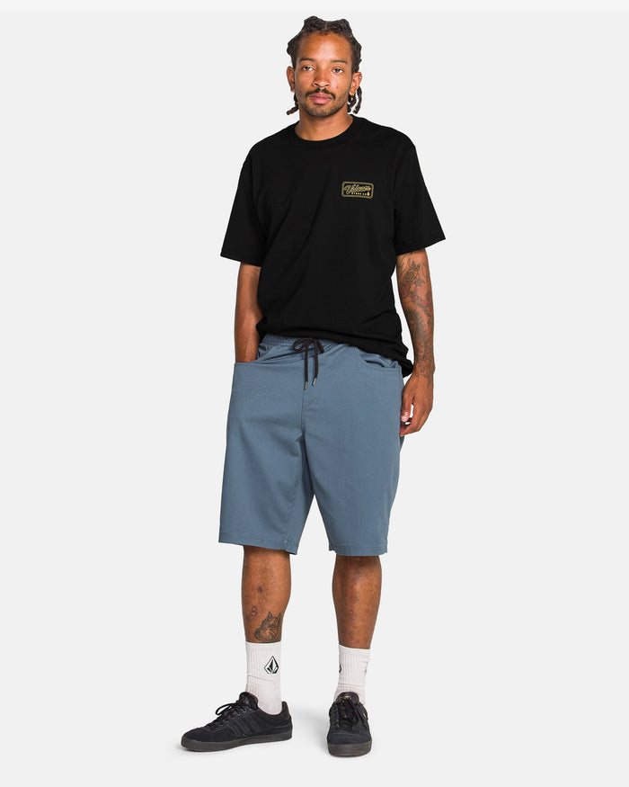 A man with braided hair faces forward, wearing the Volcom Men's Wrecking Short Sleeve Tee in black, light blue knee-length shorts, white socks, and black sneakers against a plain white background.