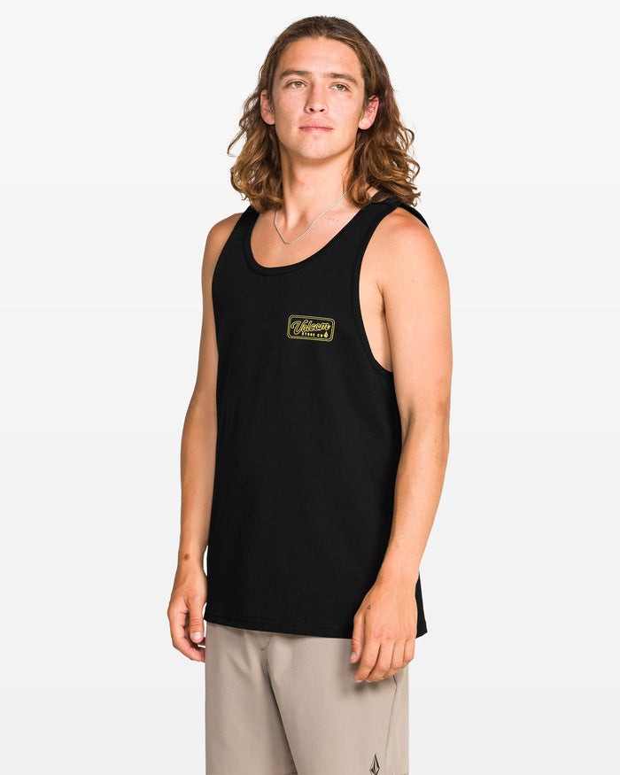 A young man with long brown hair models the Volcom Men's Wrecking Tank in a classic fit with a small yellow chest graphic, paired with beige shorts, set against a plain white background.
