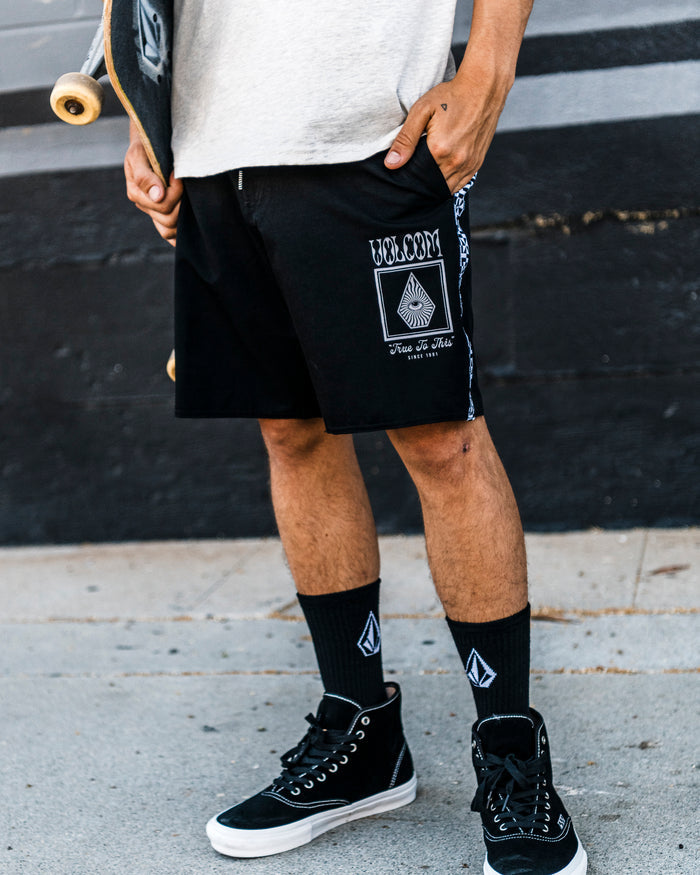 A person wearing Volcom Men's Yewster Loose Stoney black boardshorts, black socks, and black sneakers stands on a sidewalk holding a skateboard. Only the lower half of their body is visible.