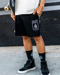 A person wearing Volcom Men's Yewster Loose Stoney black boardshorts, black socks, and black sneakers stands on a sidewalk holding a skateboard. Only the lower half of their body is visible.
