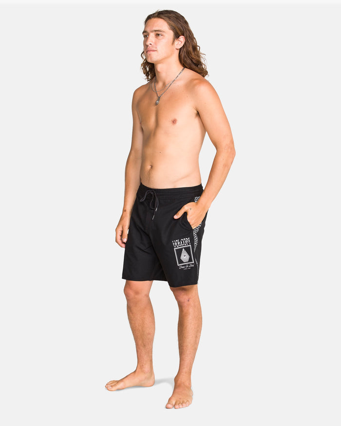A shirtless person with long hair stands barefoot, wearing Volcom Men's Yewster Loose Stoney black boardshorts featuring white graphics on the left leg and pocket, made with REPREVE recycled fibers, against a plain white background.