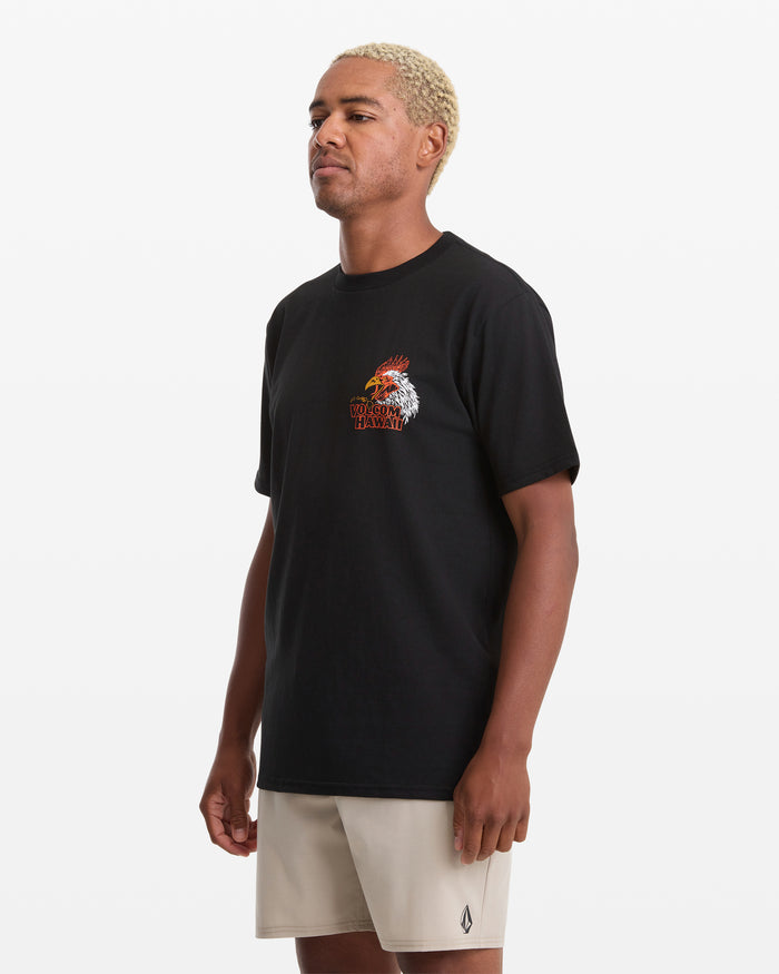 A man with short blond hair wears the Volcom Men's Yewster Rooster Short Sleeve Tee, which features a colorful graphic, paired with light beige shorts. He stands against a plain white background, looking slightly to the side.
