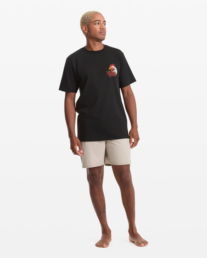A person with short bleached blond hair models the Volcom Men's Yewster Rooster Short Sleeve Tee in black and light beige shorts, standing barefoot on a plain white background and looking slightly to the side.