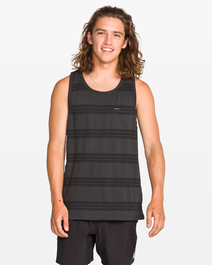 A young man with long brown hair stands and smiles at the camera against a plain white background, wearing a modern fit Men's Yewster Tank by Volcom in black and gray jacquard stripes paired with black shorts.