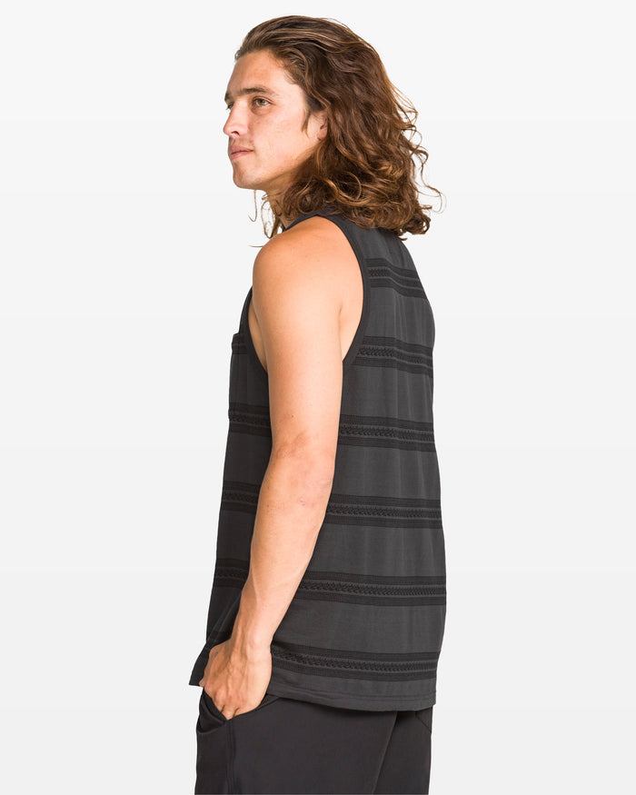 A man with long brown hair stands sideways, looking over his shoulder, wearing the Volcom Men's Yewster Tank—a modern fit, sleeveless 240gsm cotton top with jacquard stripes—paired with black pants against a plain white background.