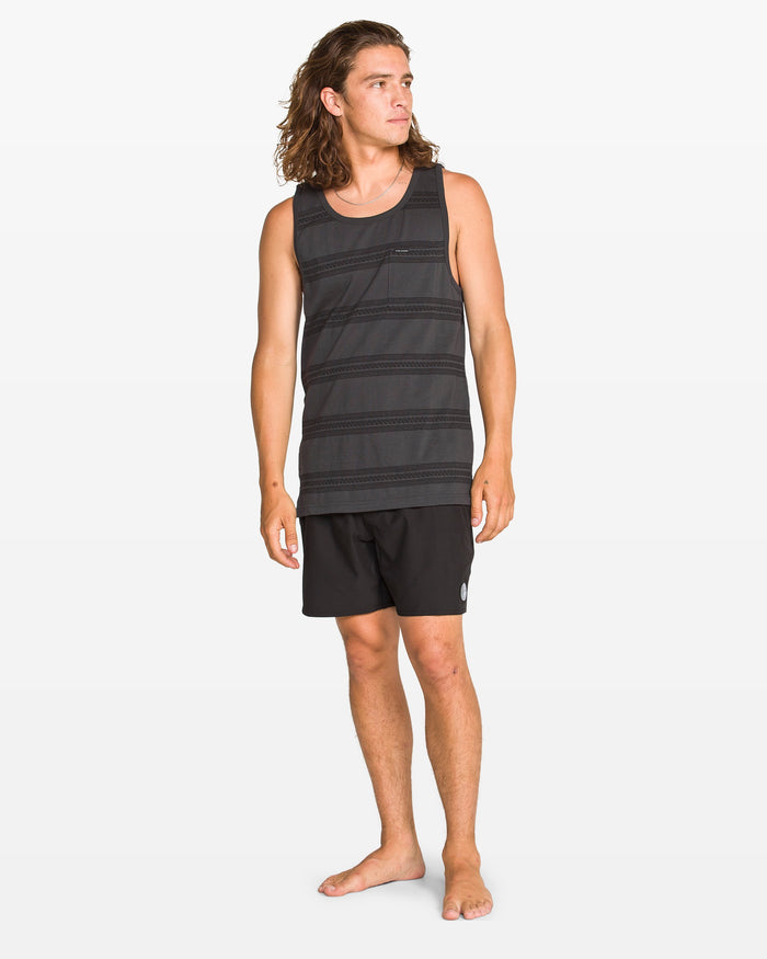 A man with long brown hair stands barefoot in Volcom's Men's Yewster Tank and black shorts, looking to his left against a plain white background.