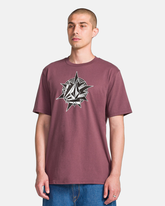A person wearing the Volcom Men's Yoink Short Sleeve Tee in maroon, featuring a black and white geometric sun and diamond graphic on the chest, paired with blue jeans, stands against a plain white background.