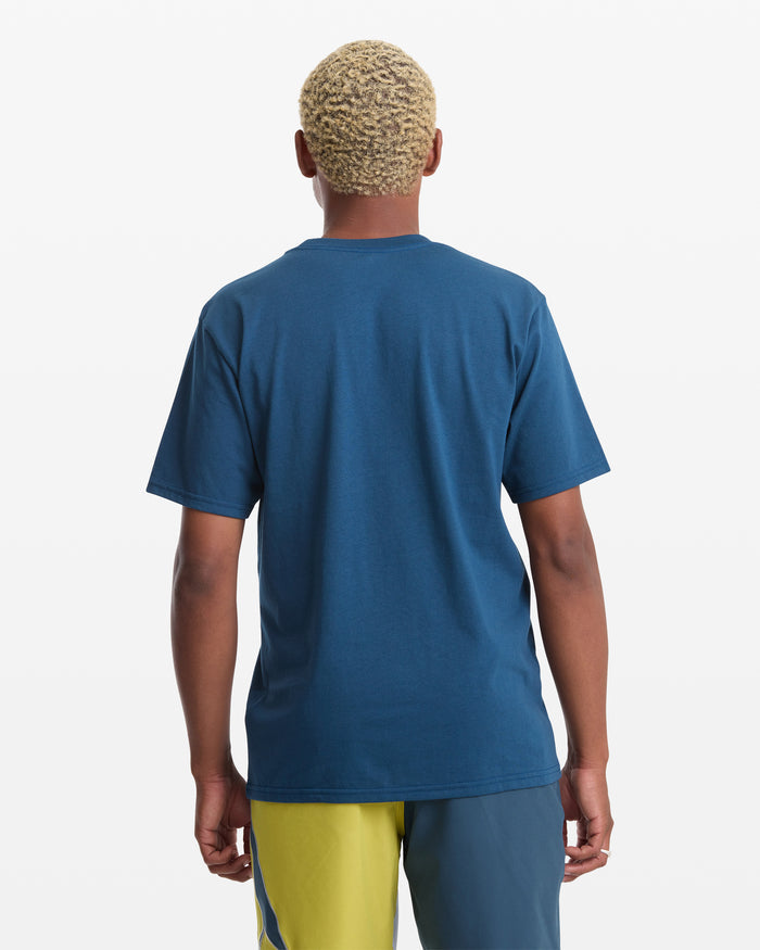 A person with short, curly blond hair is seen from behind wearing the Volcom Men's Yoink Short Sleeve Tee and color-blocked yellow and teal shorts, set against a white background.