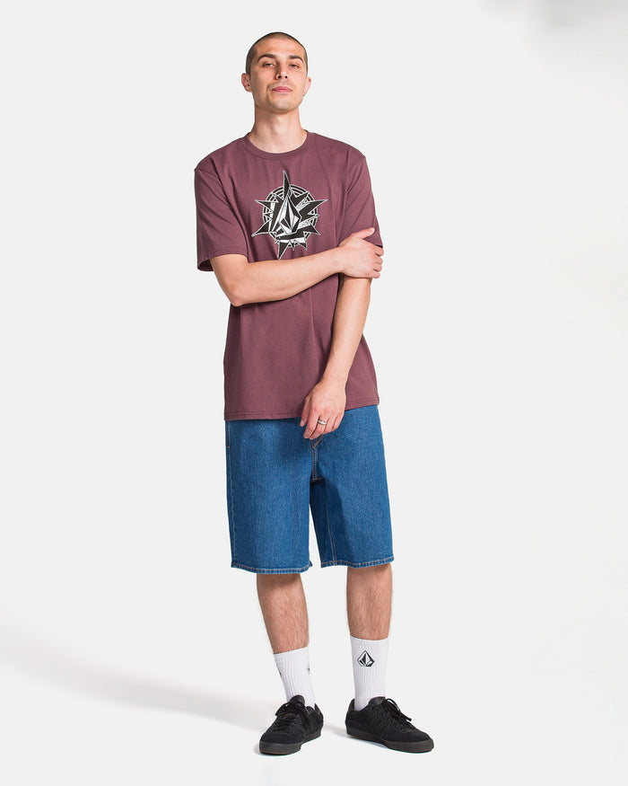 Against a white background, a person with short hair wears the Volcom Men's Yoink Short Sleeve Tee in maroon (100% cotton, classic fit, crew neck), blue denim shorts, white logo socks, and black sneakers. One hand touches their opposite arm.