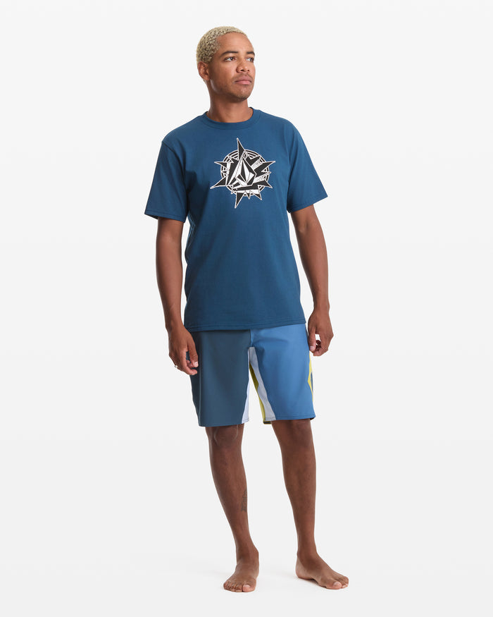 A person with short, light blonde hair stands barefoot against a white background, wearing the Volcom Men's Yoink Short Sleeve Tee in blue (100% cotton, soft-hand screen print) and blue board shorts with a yellow stripe.