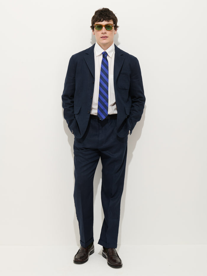 A man stands against a white background wearing the Mercer Linen Jacket In Linen, paired with a white dress shirt, blue striped tie, brown dress shoes, and green sunglasses, with his hands in his pockets.