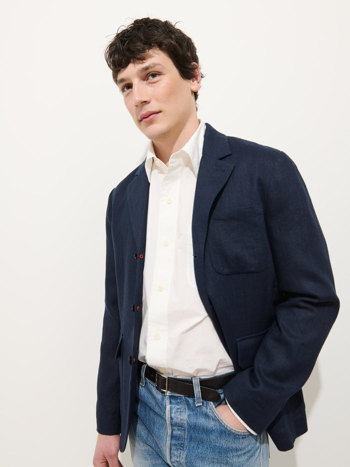 A young man poses against a white background wearing the Mercer Linen Jacket In Linen over a white button-up shirt, blue jeans, and a black belt, with one hand in his pocket.