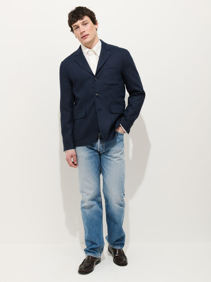 A person stands against a white background, wearing the Mercer Linen Jacket In Linen over a white shirt, light blue jeans, and dark shoes, with one hand in their pocket and a relaxed posture.