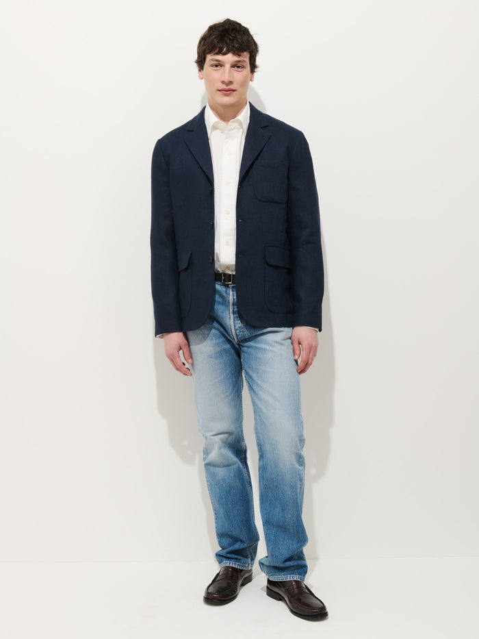 A person with short dark hair and a neutral expression stands against a white background, wearing the Mercer Linen Jacket in linen, a white shirt, blue jeans, and dark brown shoes.