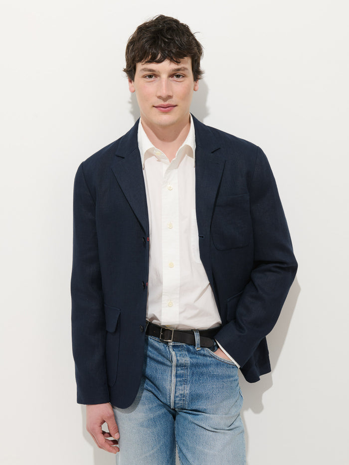 A young man with short dark hair wears the Mercer Linen Jacket in linen over a white dress shirt and blue jeans. He stands against a plain white background, one hand in his pocket, looking at the camera and smiling slightly.