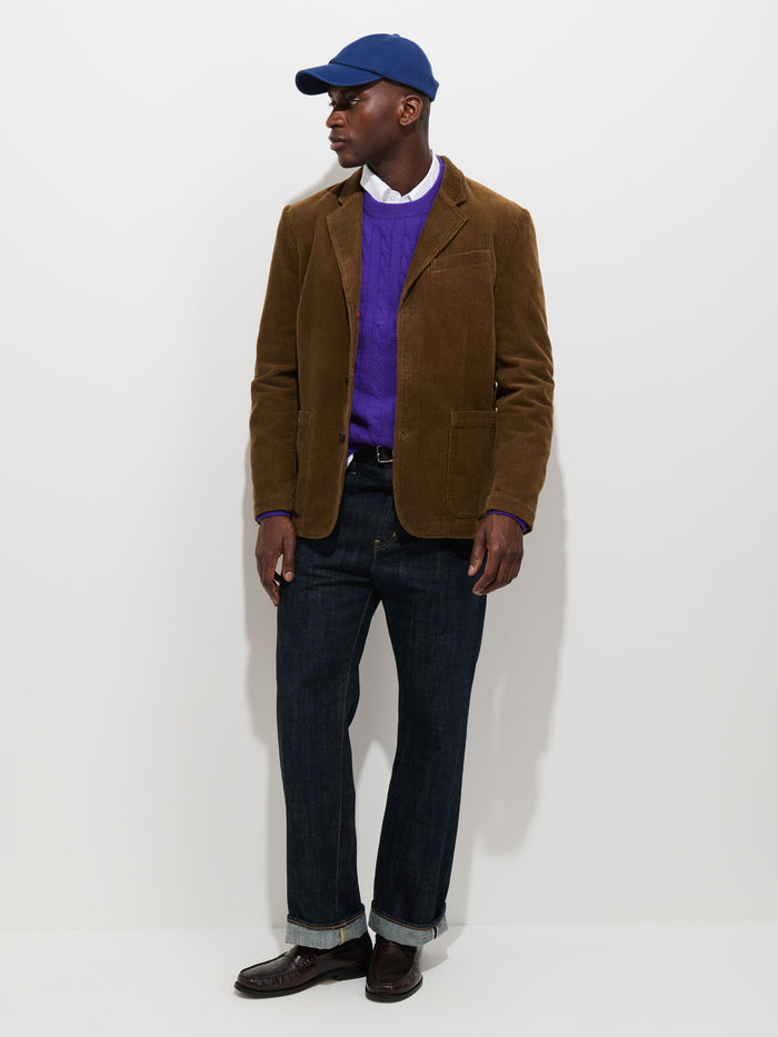 A man stands against a plain white background, looking to the side, dressed in the Mill Blazer In Corduroy—a fully lined brown cotton cord jacket—paired with a blue cap, purple sweater, white shirt, dark jeans, and brown shoes.