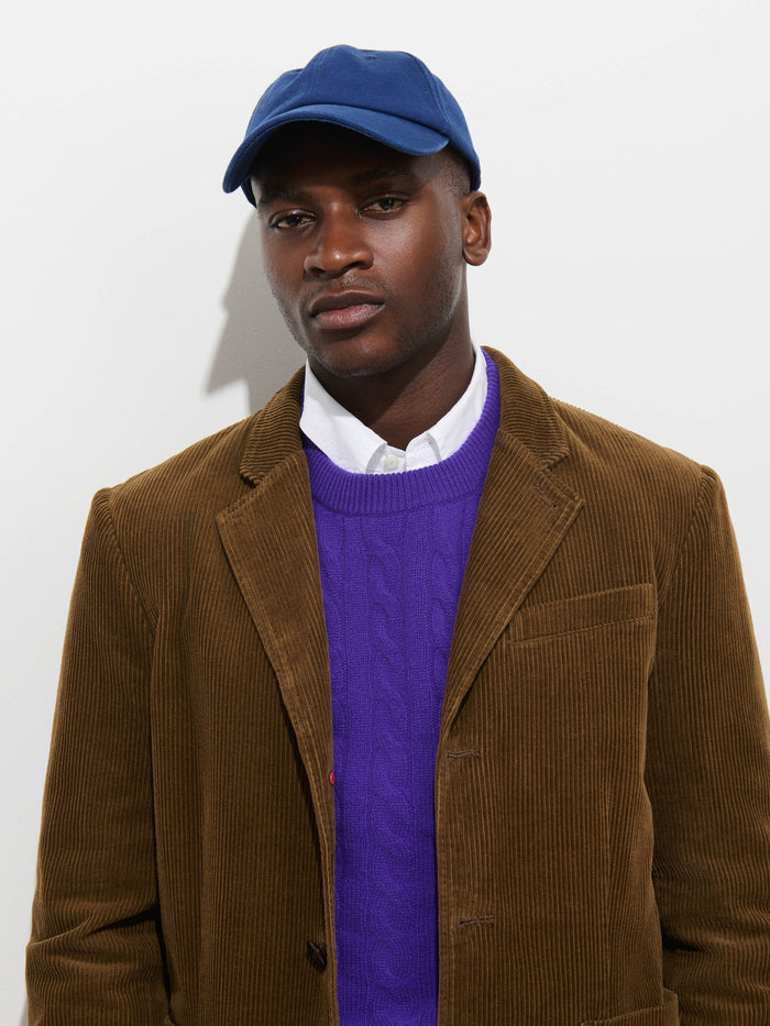 A man wearing the Mill Blazer in Corduroy stands against a plain white background, looking at the camera with a neutral expression. He pairs the brown corduroy blazer with a blue cap, white shirt, and purple sweater.