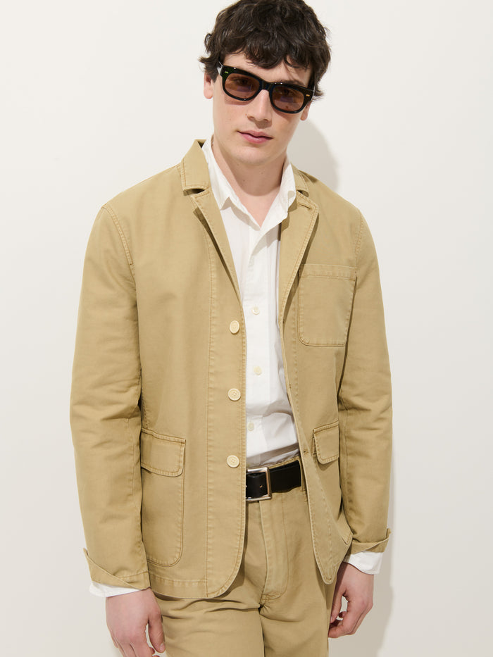 A man wears the Mill Blazer In Vintage Twill with matching pants, a white dress shirt, black belt, and dark sunglasses while standing against a plain white background.