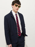 A man wears the Mill Blazer In Vintage Twill, a white dress shirt, red and navy striped tie, dark trousers, and yellow sunglasses, standing against a plain white background with one hand in his pocket.