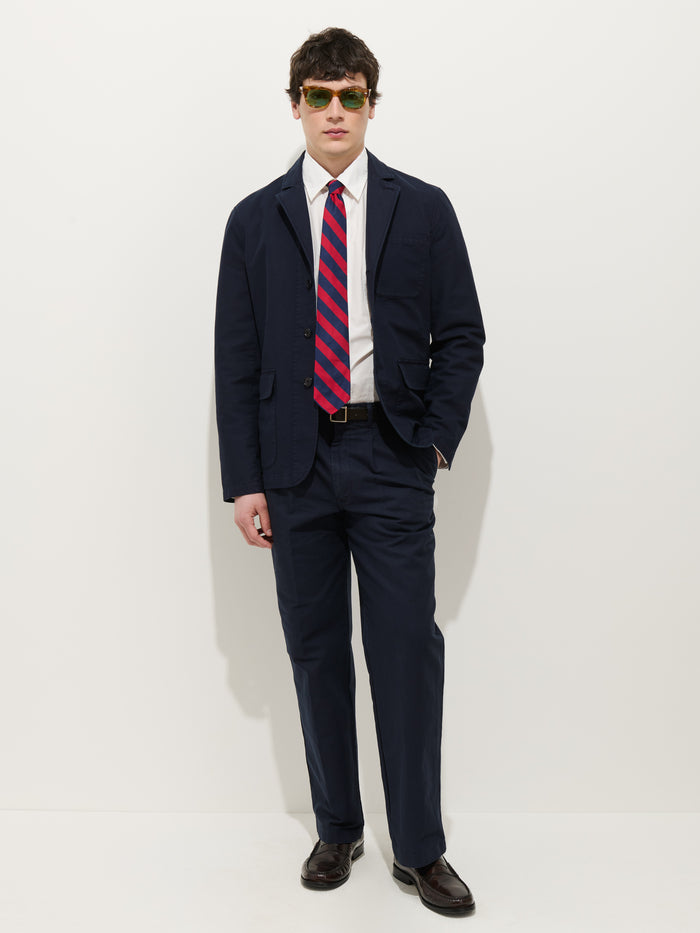 A man stands against a white background wearing a navy Mill Blazer In Vintage Twill, white dress shirt, red and navy striped tie, dark belt and shoes, and green sunglasses. One hand is in his pocket as he faces forward.