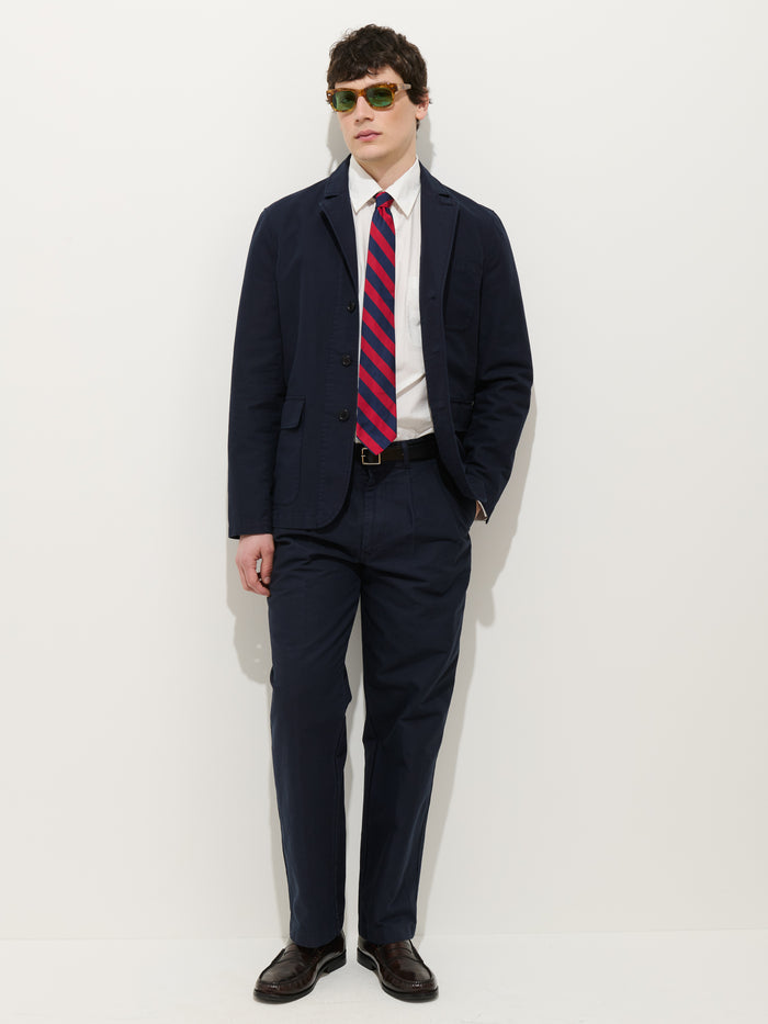 A man wearing sunglasses, the Mill Blazer In Vintage Twill in navy over a white dress shirt, a red and blue striped tie, and dark dress shoes stands against a plain white background.