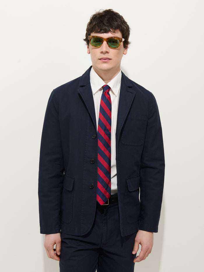A man wearing a Mill Blazer In Vintage Twill, white shirt, red and navy striped tie, dark pants, and green-tinted sunglasses stands against a plain white background.