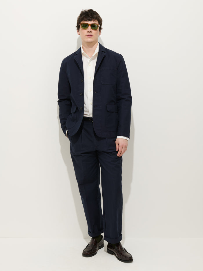 A man stands against a white background wearing the Mill Blazer in Vintage Twill, paired with a white shirt, dark dress shoes, and sunglasses. One hand is in his pocket and he poses with a relaxed posture.
